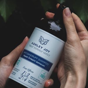 Close-up of hands holding an Ashley Joy anti-dandruff shampoo bottle with nature background.
